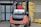 A man wearing a red vest operates a forklift in an industrial setting. The forklift appears weathered and is positioned near a large stack of yellow packaged materials labeled 'Premium'. There is a propane tank attached to the forklift, and a fire extinguisher is mounted nearby. The area is part of a warehouse or similar facility with large doors and additional pallets of materials in the background.