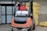 A man wearing a red vest operates a forklift in an industrial setting. The forklift appears weathered and is positioned near a large stack of yellow packaged materials labeled 'Premium'. There is a propane tank attached to the forklift, and a fire extinguisher is mounted nearby. The area is part of a warehouse or similar facility with large doors and additional pallets of materials in the background.