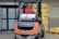 A man wearing a red vest operates a forklift in an industrial setting. The forklift appears weathered and is positioned near a large stack of yellow packaged materials labeled 'Premium'. There is a propane tank attached to the forklift, and a fire extinguisher is mounted nearby. The area is part of a warehouse or similar facility with large doors and additional pallets of materials in the background.