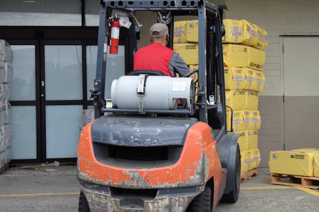 A man wearing a red vest operates a forklift in an industrial setting. The forklift appears weathered and is positioned near a large stack of yellow packaged materials labeled 'Premium'. There is a propane tank attached to the forklift, and a fire extinguisher is mounted nearby. The area is part of a warehouse or similar facility with large doors and additional pallets of materials in the background.