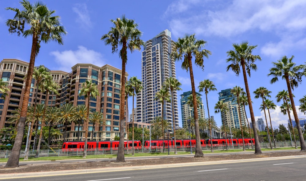 a red train traveling past tall buildings and palm trees