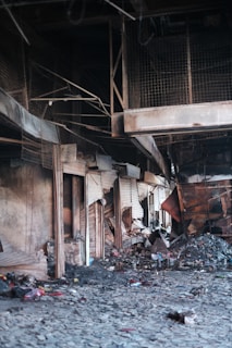 A deserted, damaged interior with debris and charred items scattered across the floor. The walls and structures are worn and partially collapsed, indicative of a recent fire or destruction. The area appears abandoned, with broken structural elements and a sense of neglect.