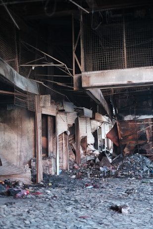 A deserted, damaged interior with debris and charred items scattered across the floor. The walls and structures are worn and partially collapsed, indicative of a recent fire or destruction. The area appears abandoned, with broken structural elements and a sense of neglect.