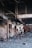 A deserted, damaged interior with debris and charred items scattered across the floor. The walls and structures are worn and partially collapsed, indicative of a recent fire or destruction. The area appears abandoned, with broken structural elements and a sense of neglect.