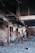 A deserted, damaged interior with debris and charred items scattered across the floor. The walls and structures are worn and partially collapsed, indicative of a recent fire or destruction. The area appears abandoned, with broken structural elements and a sense of neglect.