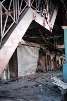 An indoor setting with visible damage and destruction. There are multiple closed, metal roll-up doors, and debris scattered across the floor including broken objects and trash. The area appears abandoned and possibly affected by fire.