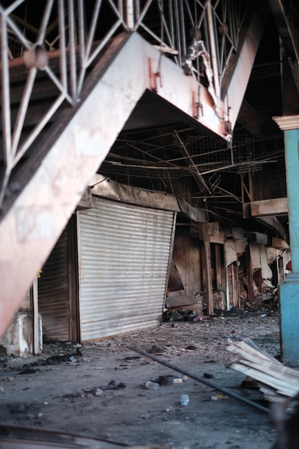 An indoor setting with visible damage and destruction. There are multiple closed, metal roll-up doors, and debris scattered across the floor including broken objects and trash. The area appears abandoned and possibly affected by fire.
