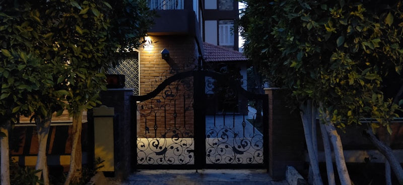 A wrought iron gate with intricate designs is illuminated by a warm light, casting shadows on the paved ground. Tall greenery frames the gate on both sides, while a brick wall and house with a tiled roof and shuttered windows stand in the background. The scene appears to be at dusk.