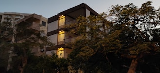A modern residential building with warm lighting at sunset, surrounded by green trees.