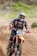 A motorcyclist wearing protective gear, including a helmet and goggles, rides a dirt bike at speed on a dirt track. The number 639 is visible on the front of the bike. The rider is surrounded by dirt and dust, with a blurred background of greenery.