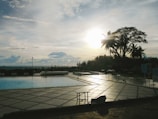 A swimming pool area with a partially cloudy sky at sunset or sunrise. The sun is low, casting long shadows across the pool deck. Tall palm trees are silhouetted against the sky, and the pool water reflects the soft golden light. Lounge chairs and tables are placed around the pool, suggesting a relaxing environment.
