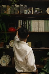 A person in a white shirt is reaching up to a shelf filled with various books and magazines, including titles like Vogue and Kinfolk. The shelf is adorned with a small plant, a red teapot, decorative plates, and a candle. The setting has an eclectic and vintage vibe.