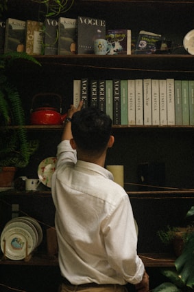 A person in a white shirt is reaching up to a shelf filled with various books and magazines, including titles like Vogue and Kinfolk. The shelf is adorned with a small plant, a red teapot, decorative plates, and a candle. The setting has an eclectic and vintage vibe.