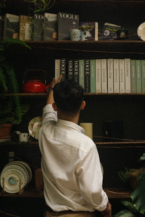 A person in a white shirt is reaching up to a shelf filled with various books and magazines, including titles like Vogue and Kinfolk. The shelf is adorned with a small plant, a red teapot, decorative plates, and a candle. The setting has an eclectic and vintage vibe.