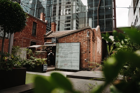 A quaint brick building with a sign reading 'Little Lon' and 'Gin Bar' framed by urban high-rises in the background. The entrance is adorned with green foliage and plants, creating an inviting atmosphere.