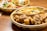 A close-up of a meal set in a plastic tray on a wooden table. The tray contains fried chicken pieces, French fries, and a small portion of mixed vegetables. Another tray in the background contains rice, more vegetables, and breaded items.