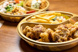 A close-up of a meal set in a plastic tray on a wooden table. The tray contains fried chicken pieces, French fries, and a small portion of mixed vegetables. Another tray in the background contains rice, more vegetables, and breaded items.