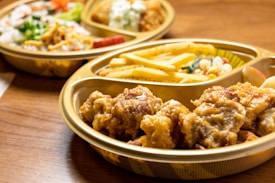 A close-up of a meal set in a plastic tray on a wooden table. The tray contains fried chicken pieces, French fries, and a small portion of mixed vegetables. Another tray in the background contains rice, more vegetables, and breaded items.
