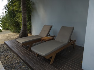Two lounge chairs with beige cushions are placed next to a wooden side table. They are situated on a wooden deck surrounded by pebbles and adjacent to a sandy area with tropical plants and a palm tree. A light blue wall adds a serene backdrop to the setting.