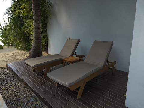 Two lounge chairs with beige cushions are placed next to a wooden side table. They are situated on a wooden deck surrounded by pebbles and adjacent to a sandy area with tropical plants and a palm tree. A light blue wall adds a serene backdrop to the setting.