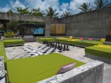 A spacious outdoor lounge area featuring several large green cushioned seating areas arranged around a giant chessboard. The chess pieces are oversized and made of black and white materials. The setting is surrounded by concrete walls and has tropical plants and palm trees visible in the background, with a vibrant blue sky.