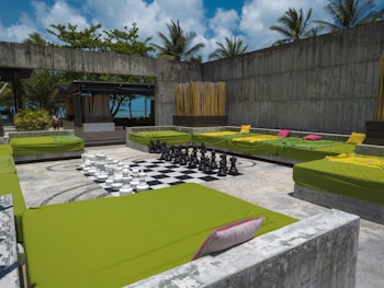 A spacious outdoor lounge area featuring several large green cushioned seating areas arranged around a giant chessboard. The chess pieces are oversized and made of black and white materials. The setting is surrounded by concrete walls and has tropical plants and palm trees visible in the background, with a vibrant blue sky.