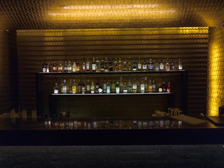 A dimly lit bar area with a backlit shelf displaying an array of liquor bottles neatly arranged. The wall behind has a textured, gold-toned appearance, adding a luxurious and warm ambiance to the setting. A dark counter in the foreground reflects some of the bottles, and a few glasses and small items are placed on it.