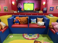 A brightly colored room features a red and blue seating arrangement with colorful cushions that have patterns like flowers and rainbows. Various plush toys, including a bear and a pink character, occupy the seats. The backdrop includes a red and pink wall with houndstooth patterns, framed pictures, and a TV screen displaying a cartoon with a bright green background. A retro telephone and other decor items are on display, while a vibrant rug with floral patterns lies on the floor.