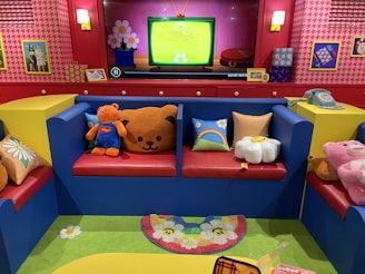 A brightly colored room features a red and blue seating arrangement with colorful cushions that have patterns like flowers and rainbows. Various plush toys, including a bear and a pink character, occupy the seats. The backdrop includes a red and pink wall with houndstooth patterns, framed pictures, and a TV screen displaying a cartoon with a bright green background. A retro telephone and other decor items are on display, while a vibrant rug with floral patterns lies on the floor.