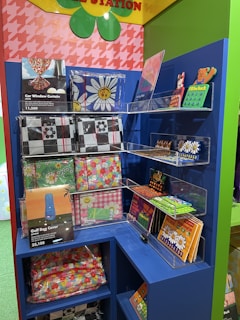 A colorful display shelf features various patterned items, including car window curtains and golf bag covers, with floral and checkered designs. The top of the display has green and yellow decorative accents with a pink and red houndstooth pattern in the background.