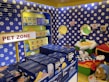 A colorful pet store section with a vibrant blue and yellow floral theme. Shelves are filled with pet party kits, grooming products, and toys. A plush cushion shaped like a flower with a smiley face is placed in the corner along with stuffed animals resembling dogs and lions. The area is labeled as 'Pet Zone' with a prominent sign.