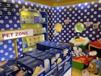A colorful pet store section with a vibrant blue and yellow floral theme. Shelves are filled with pet party kits, grooming products, and toys. A plush cushion shaped like a flower with a smiley face is placed in the corner along with stuffed animals resembling dogs and lions. The area is labeled as 'Pet Zone' with a prominent sign.