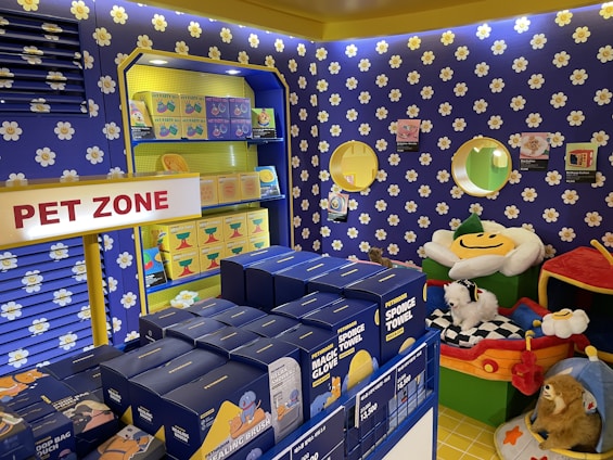A colorful pet store section with a vibrant blue and yellow floral theme. Shelves are filled with pet party kits, grooming products, and toys. A plush cushion shaped like a flower with a smiley face is placed in the corner along with stuffed animals resembling dogs and lions. The area is labeled as 'Pet Zone' with a prominent sign.