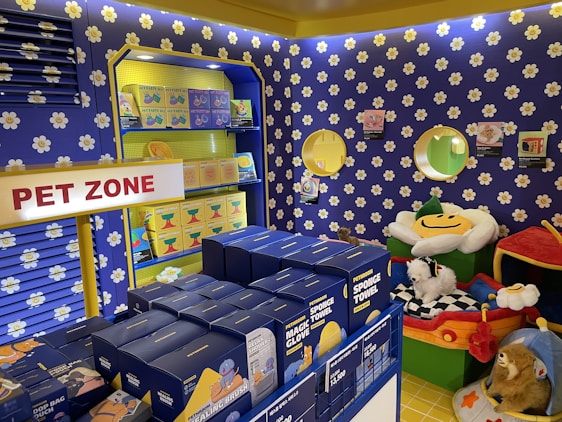 A colorful pet store section with a vibrant blue and yellow floral theme. Shelves are filled with pet party kits, grooming products, and toys. A plush cushion shaped like a flower with a smiley face is placed in the corner along with stuffed animals resembling dogs and lions. The area is labeled as 'Pet Zone' with a prominent sign.