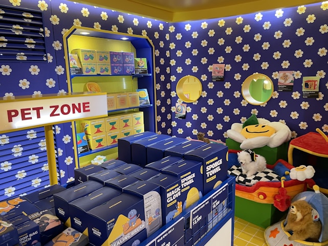 A colorful pet store section with a vibrant blue and yellow floral theme. Shelves are filled with pet party kits, grooming products, and toys. A plush cushion shaped like a flower with a smiley face is placed in the corner along with stuffed animals resembling dogs and lions. The area is labeled as 'Pet Zone' with a prominent sign.