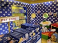A colorful pet store section with a vibrant blue and yellow floral theme. Shelves are filled with pet party kits, grooming products, and toys. A plush cushion shaped like a flower with a smiley face is placed in the corner along with stuffed animals resembling dogs and lions. The area is labeled as 'Pet Zone' with a prominent sign.