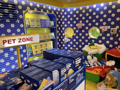 A colorful pet store section with a vibrant blue and yellow floral theme. Shelves are filled with pet party kits, grooming products, and toys. A plush cushion shaped like a flower with a smiley face is placed in the corner along with stuffed animals resembling dogs and lions. The area is labeled as 'Pet Zone' with a prominent sign.