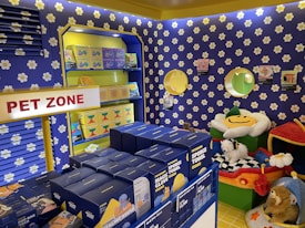 A colorful pet store section with a vibrant blue and yellow floral theme. Shelves are filled with pet party kits, grooming products, and toys. A plush cushion shaped like a flower with a smiley face is placed in the corner along with stuffed animals resembling dogs and lions. The area is labeled as 'Pet Zone' with a prominent sign.