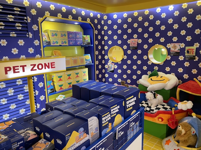 A colorful pet store section with a vibrant blue and yellow floral theme. Shelves are filled with pet party kits, grooming products, and toys. A plush cushion shaped like a flower with a smiley face is placed in the corner along with stuffed animals resembling dogs and lions. The area is labeled as 'Pet Zone' with a prominent sign.