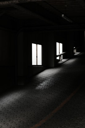 A dimly lit parking garage with a sloped, textured floor leading upwards, illuminated by a series of rectangular windows casting natural light inside.