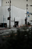 A construction site covered with white sheets, situated at a street corner with visible road signs and traffic lights. Two individuals wearing helmets stand near the entrance. The scene includes some parked and passing vehicles, and the background is a large building with small windows.