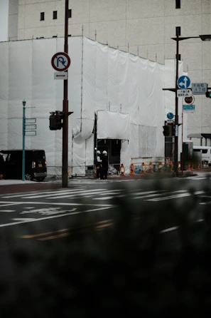 A construction site covered with white sheets, situated at a street corner with visible road signs and traffic lights. Two individuals wearing helmets stand near the entrance. The scene includes some parked and passing vehicles, and the background is a large building with small windows.