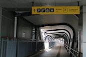 A modern indoor walkway with a geometric ceiling structure and a pathway leading into the distance. The walkway is enclosed by metal railings and features a yellow directional sign with icons for exit, stairs, and other facilities.
