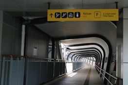 A modern indoor walkway with a geometric ceiling structure and a pathway leading into the distance. The walkway is enclosed by metal railings and features a yellow directional sign with icons for exit, stairs, and other facilities.