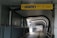 A modern indoor walkway with a geometric ceiling structure and a pathway leading into the distance. The walkway is enclosed by metal railings and features a yellow directional sign with icons for exit, stairs, and other facilities.