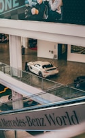A sleek white sports car is displayed indoors on a wooden floor, surrounded by modern architecture with large glass railings. Above, large advertising graphics show people, while a Mercedes-Benz World sign is visible.