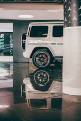 A white luxury SUV is parked in an indoor showroom with shiny black flooring. The vehicle's smooth exterior and sleek design are complemented by polished black wheels. Reflection of the car is visible on the glossy floor, enhancing the elegance of the setting. A partial view of a column with a star pattern is seen on the right, contributing to the upscale atmosphere.