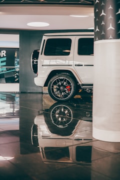 A white luxury SUV is parked in an indoor showroom with shiny black flooring. The vehicle's smooth exterior and sleek design are complemented by polished black wheels. Reflection of the car is visible on the glossy floor, enhancing the elegance of the setting. A partial view of a column with a star pattern is seen on the right, contributing to the upscale atmosphere.