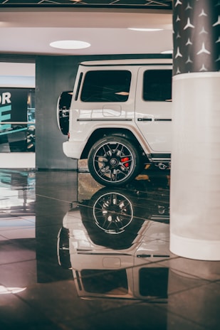 A white luxury SUV is parked in an indoor showroom with shiny black flooring. The vehicle's smooth exterior and sleek design are complemented by polished black wheels. Reflection of the car is visible on the glossy floor, enhancing the elegance of the setting. A partial view of a column with a star pattern is seen on the right, contributing to the upscale atmosphere.