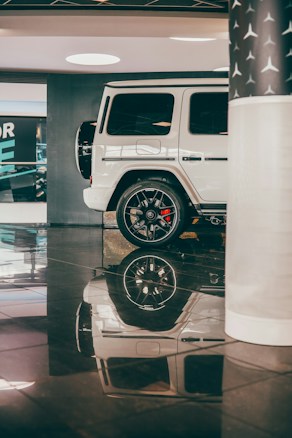 A white luxury SUV is parked in an indoor showroom with shiny black flooring. The vehicle's smooth exterior and sleek design are complemented by polished black wheels. Reflection of the car is visible on the glossy floor, enhancing the elegance of the setting. A partial view of a column with a star pattern is seen on the right, contributing to the upscale atmosphere.